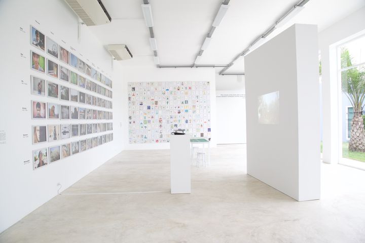 The installation with no one in it: a photo wall, the wall of aliens at the back, a free-standing white wall and a plinth, Thaillywood